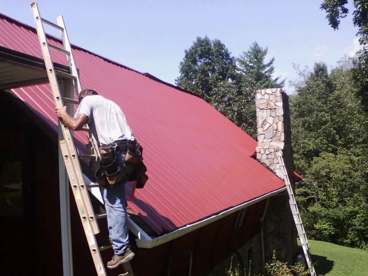 Roofer on a ladder beside a red metal roof and stone chimney — Soffit & Fascia Repair by Roofing Brothers in Woodward